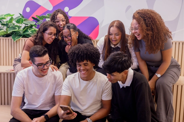 Imagem de jovens sentados juntos e sorrindo enquanto olham pra um celular.